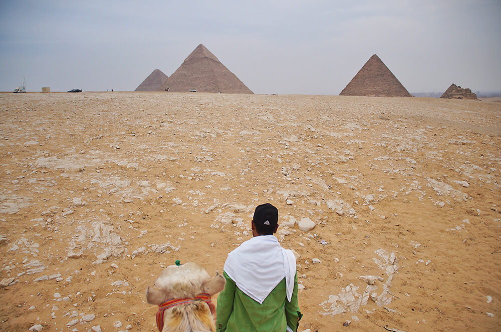 騎乘在駱駝上方的微貓視角，黃色沙漠，金字塔，白色岩石，駱駝與商人。